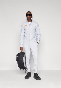 Šviesiai pilka zip-up striukė su kapišonu, juodais akcentais ir logotipu; atitinkančios sportinės kelnės; juodas kuprinė; juodi batai; didelės saulės akiniai.