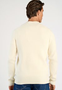 Gräddfärgad ribbad stickad tröja med långa ärmar, rundad halsringning och sidoventilationsdetalj, i kombination med blå denimjeans.