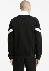 Fekete sportos dzseki, fehér csíkkal a vállaknál, bordázott mandzsettákkal és magas gallérral, a kényelem és a mozgás érdekében tervezve.