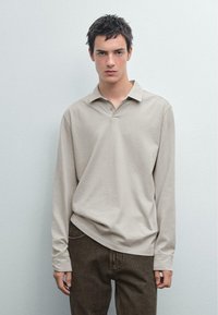 Beige polo shirt met lange mouwen en een kraag, heeft een comfortabele pasvorm en een ronde onderrand, gecombineerd met donkerbruine jeans. Zachte textuur.
