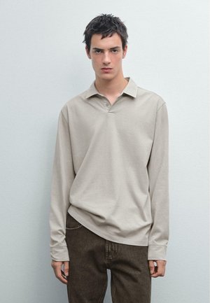 Long-sleeve beige polo shirt with a collar, featuring a relaxed fit and rounded hem, paired with dark brown jeans. Smooth texture.