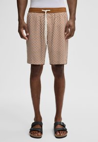 Uomo che indossa pantaloni corti a righe marroni e bianchi con coulisse, in piedi a piedi nudi con sandali neri con fibbia su uno sfondo semplice.