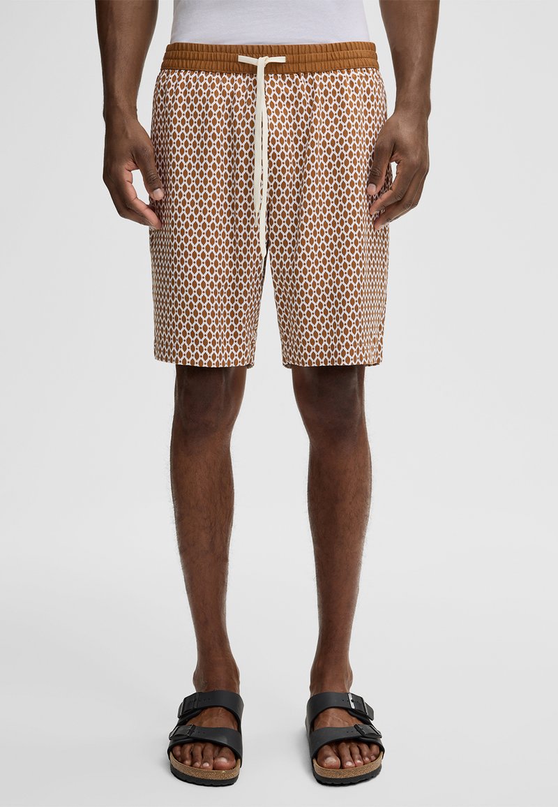 Uomo che indossa pantaloni corti a righe marroni e bianchi con coulisse, in piedi a piedi nudi con sandali neri con fibbia su uno sfondo semplice.