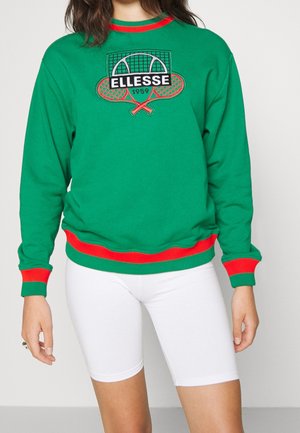 Zielona bluza z czerwonym obszyciem i logo tenisa z napisem "ELLESSE 1959,", noszona z białymi dopasowanymi spodenkami rowerowymi.