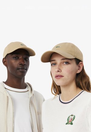 Beige baseballcaps laget av bomull med bøyd brimmel og brodert logo. To modeller har på seg uformelle klær med kontrasterende teksturer og farger.