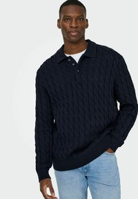 Marineblaues Kabelstrick-Pullover mit Polo-Kragen, ausgestattet mit drei Knöpfen, gerippten Bündchen und einem strukturierten Design. Über einem weißen Hemd getragen.