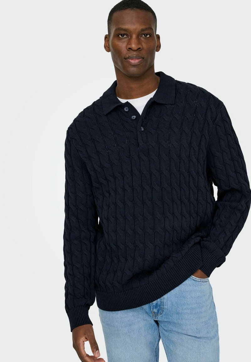 Marineblaues Kabelstrick-Pullover mit Polo-Kragen, ausgestattet mit drei Knöpfen, gerippten Bündchen und einem strukturierten Design. Über einem weißen Hemd getragen.