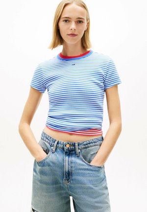 Młoda kobieta z krótkimi blond włosami, ubrana w niebiesko-biały, paskowany krótki T-shirt i jasnoniebieskie, wysokie szorty jeansowe, z rękami w kieszeniach.