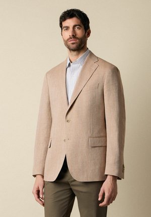 Mężczyzna ubrany w beżową marynarkę na dwa guziki, jasnoniebieską koszulę w kratę oraz oliwkowo-zielone spodnie, stojący na jednolitym beżowym tle.