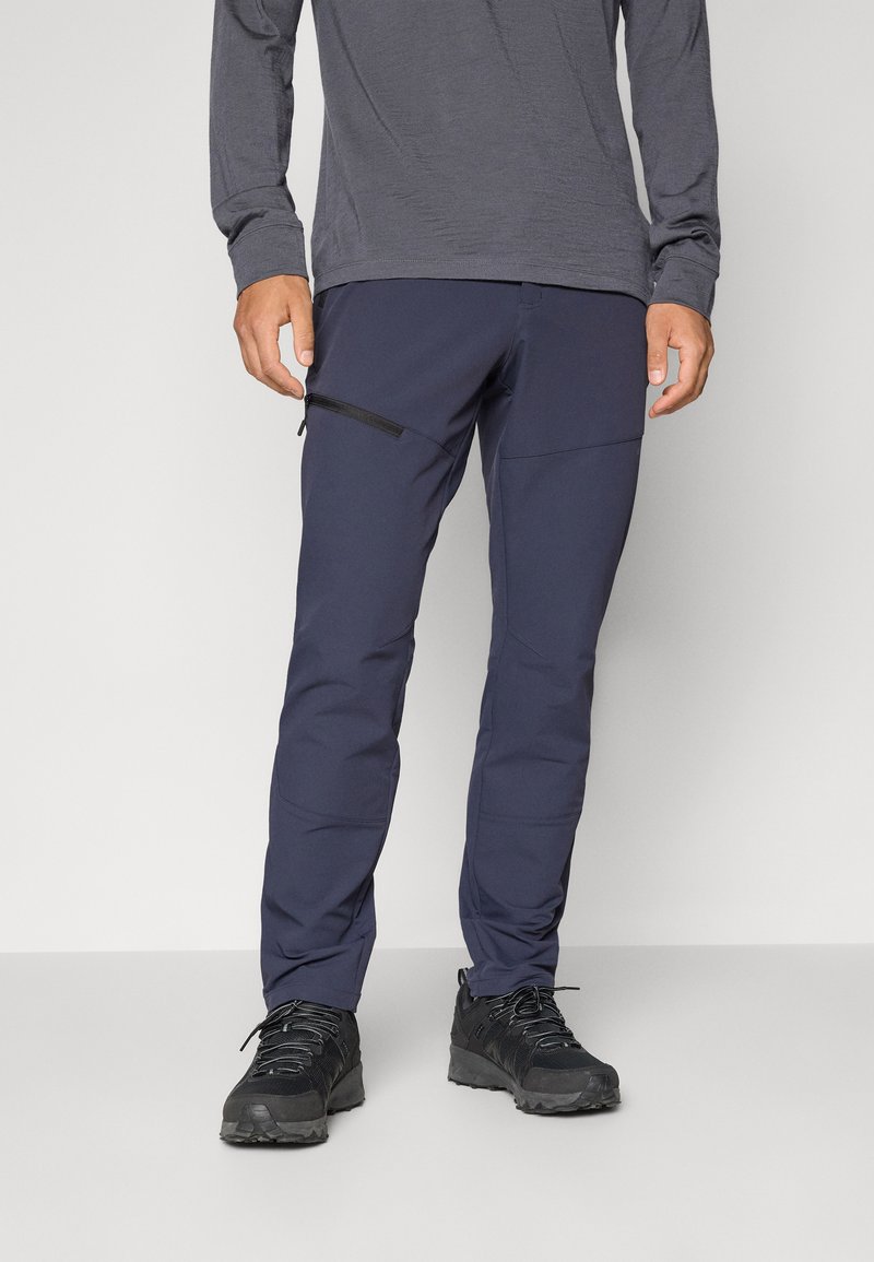 Sötétkék, slim fit szabású kültéri nadrág, cipzáras zsebre, puha tapintású anyaggal és megerősített térdrésszel. Fekete lábbeli látható alul.