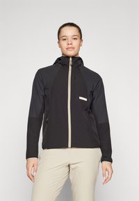 Veste à capuche noire avec une fermeture éclair beige, dotée d'une poche poitrine zippée et d'un design de panneaux contrastés. Matériau lisse et léger.