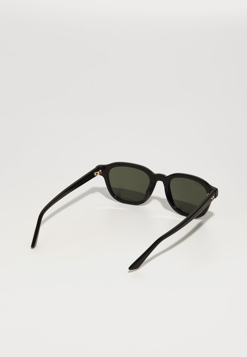 Lunettes de soleil noires avec une monture arrondie, des verres vert foncé et une texture plastique lisse. Présentent des accents dorés sur les charnières.