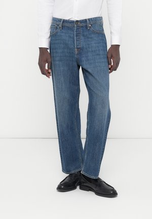 Homme portant un jean droit bleu moyen, des chaussures en cuir noires et une chemise blanche boutonnée, debout devant un fond uni.