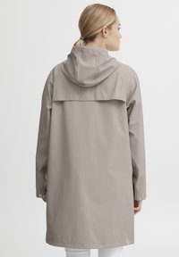 Lys beige regnfrakk med hette, med en lengre lengde, trykknapplukking ved ermene og ventilerte bakdesign for god luftsirkulasjon og komfort.