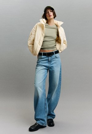 Jeune femme portant une doudoune beige, un haut court vert, un jean bleu ample, une ceinture noire, des chaussures noires et une casquette verte sur un fond gris.