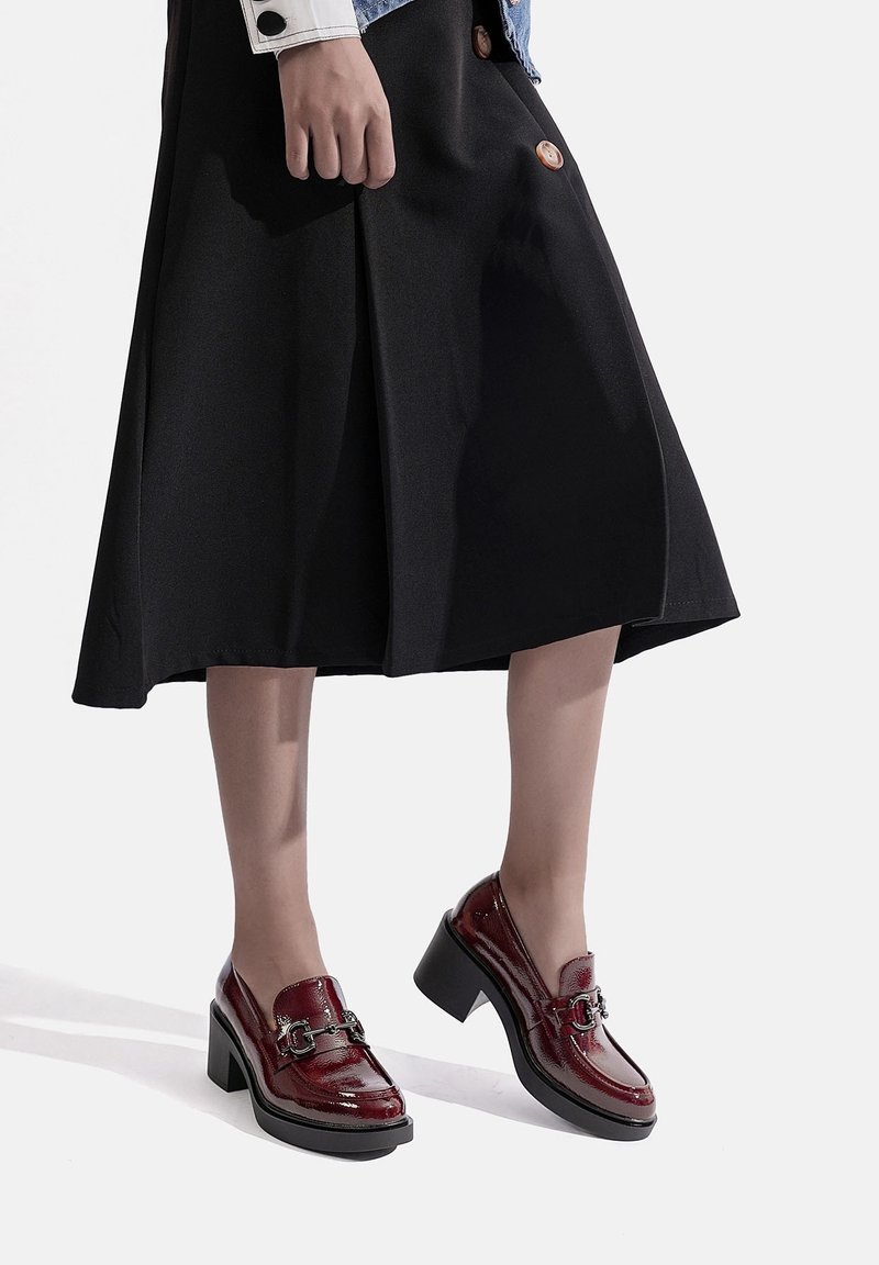 Femme portant une jupe midi noire et des mocassins à talons bordeaux brillants avec un détail de boucle en métal, debout devant un fond blanc.