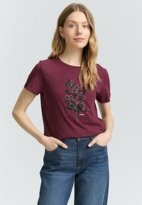 Femme aux longs cheveux blonds portant un t-shirt bordeaux avec un dessin floral en noir et blanc et un jean bleu, les mains dans les poches, sur un fond neutre.