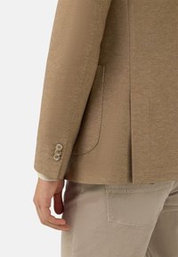 Beżowa marynarka w jodełkę o dopasowanym kroju, z czterema guzikami na mankiecie i pojedynczym rozcięciem z tyłu. W połączeniu z jasnobeżowymi spodniami z sztruksu.