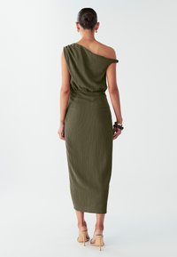 Olijfgroene, asymmetrische jurk met één schouder, gemaakt van gestructureerde stof, aansluitende taille en een zoom tot op de knie. Heeft een geplooid detail bij de schouder en een gladde, gestroomlijnde vorm.