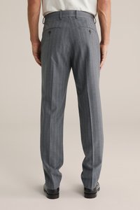 Mężczyzna ubrany w szare spodnie garniturowe w prążki i czarne eleganckie buty, stoi z rękami swobodnie opuszczonymi wzdłuż ciała na tle jednolitego tła.