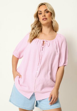 Kvinde iført en lyserød tekstureret bluse med knapper og bindebånd ved halsudskæringen, kombineret med lyseblå denimshorts.