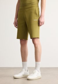 Maslinaste zelene šortse s mekom teksturom i elastičnom trakicom; sadrže Calvin Klein logo u bijeloj boji s lijeve strane. Upareni s bijelim tenisicama.