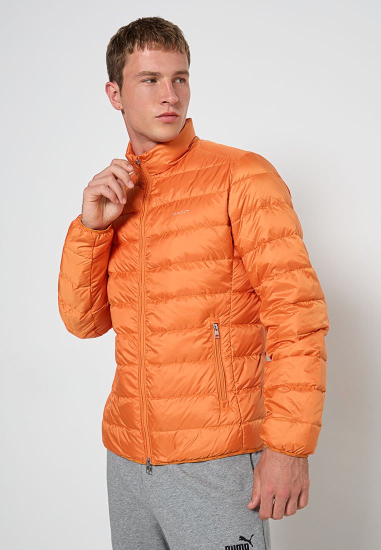 Orangene gepolsterte Jacke mit hohem Kragen, horizontalen Nähten, Reißverschlusstaschen und leichtem Material. GANT-Logo auf der Brust.
