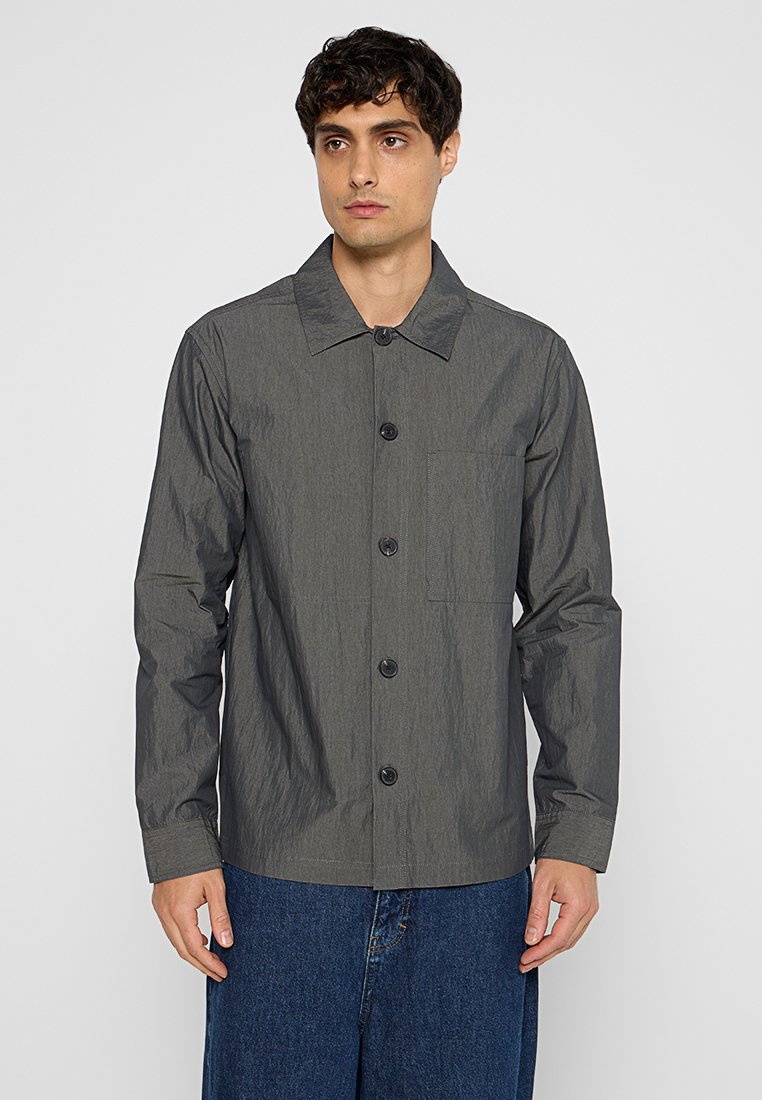 Mężczyzna w ciemnoszarej, długiej koszuli z guzikami, z kieszonką na piersi, oraz w luźnych niebieskich dżinsach, stoi na tle gładkiej ściany.