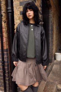 Giacca bomber nera in ecopelle con maniche a palloncino, abbinata a una t-shirt a righe verdi e bianche e a una gonna plissettata marrone. Calze alte grigie fino al ginocchio.