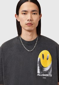 Sort t-shirt med et gult smiley face grafisk design og "ALLSAINTS" logo. Bomuldsmateriale, korte ærmer, afslappet pasform, med sølv kæden halskæde.