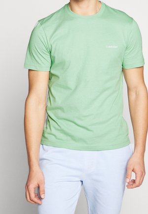 Muž nosící světle zelené tričko Calvin Klein s krátkým rukávem a světlé modré kalhoty na jednobarevném pozadí.