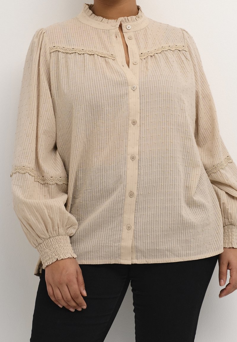 Blouse beige à manches longues avec fermeture boutonnée, texture plissée et accents en dentelle sur les épaules et les poignets. Tissu lisse avec des points subtils.