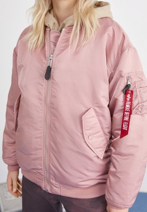 Osoba nosząca różową kurtkę typu bomber z beżową bluzą z kapturem pod spód, z czerwoną metką "Remove Before Flight" na zamku kieszeni na rękawie.