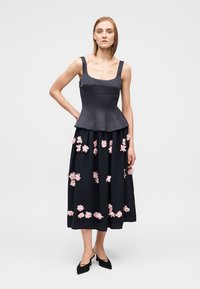 Πεπιεσμένη μπλούζα από σκουρόχρωμο γκρι ύφασμα, συνδυασμένη με μαύρη φούστα διακοσμημένη με ροζ κεντήματα λουλουδιών, με γραμμή που ανοίγει και μαύρα μυτερά παπούτσια.