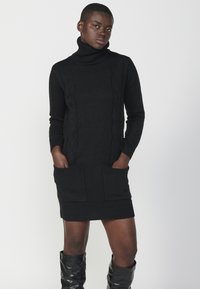 Abito maglione nero in maglia con collo alto, dettagli a coste e due tasche frontali. Presenta un design a cables testurizzato.