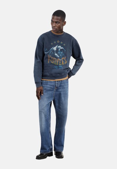 Marineblauwe sweatshirt met een grafisch panterontwerp en het woord "KOOPLES," gecombineerd met lichtblauwe straight-leg jeans en zwarte schoenen.