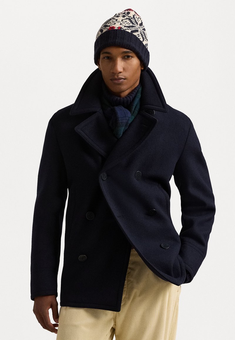 Péacoat en laine bleu marine avec un design croisé, de grands revers et des boutons noirs. Associé à un bonnet en tricot à motif et une écharpe à carreaux.
