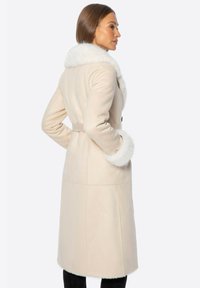 Beżowy płaszcz z futrzanym kołnierzem i mankietami, z paskiem w talii i prostym krojem. Gładka tekstura z minimalnymi detalami szwów.