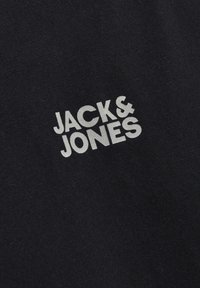 Sort bomuldsstof med teksten "JACK & JONES" i lys grå, hvor skrifttypen er ren og moderne, placeret diagonalt over materialet.