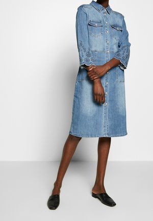 Femme portant une robe en denim bleu longueur genou avec des manches brodées et une fermeture boutonnée à l'avant, associée à des chaussures noires sans lacets.
