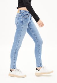 Jean skinny en denim bleu clair, avec une taille mi-haute, cinq poches et une pièce en cuir beige à l'arrière. Des baskets blanches complètent le look.
