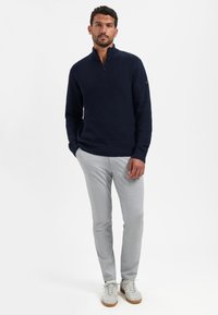 Man staat in een marineblauwe half-zip gebreide trui, lichtgrijze broek en witte sneakers voor een plain witte achtergrond.