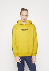 Yellow cotton hoodie with a front pocket, black logo, and side stripe detailing. Features a drawstring hood and relaxed fit.