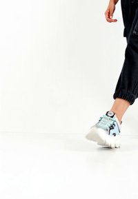 Inimene, kes kannab musti pükse ja helesiniseid-valgeid tossusid, kõnnib valgel põrandal valge tausta ees.