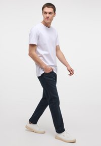 Bărbat care merge purtând un tricou alb cu mâneci scurte, pantaloni bleumarin și adidași albi pe un fundal simplu și deschis la culoare.