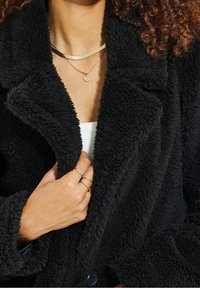 Cappotto in peluche nero con finitura testurizzata, ampio colletto e chiusura con bottoni. Collane sovrapposte e anelli completano l'outfit.