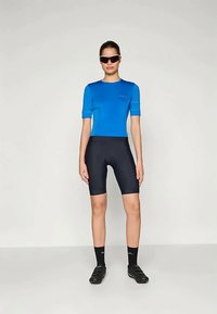 Naine, kandes sinist jalgratturi särki, musti jalgrattapükse, musti sokke, musti jalgrattakingi ja prille, seisab ühtlasel taustal.