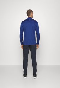 Tmavě modrá košile s dlouhým rukávem a vysokým límcem, v kombinaci s tmavými kalhotami a černými sportovními botami, z pohledu zezadu na bílém pozadí.