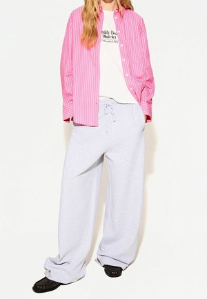 Person trägt weite weiße Jogginghosen, schwarze Schuhe, ein weißes T-Shirt mit Grafik und ein aufgeknöpftes, rosa gestreiftes langärmeliges Hemd.