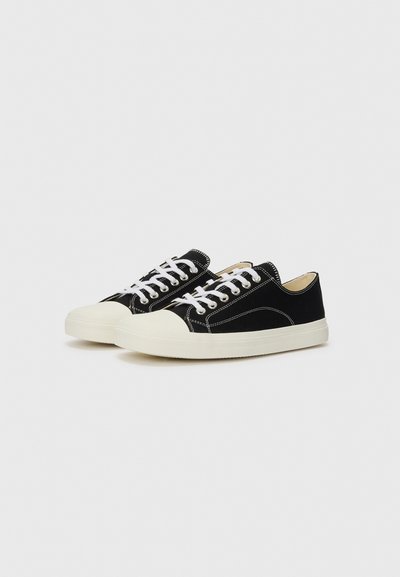 Svarta kanvas sneakers med vita snörningar, krämfärgad gummikap och kontrasterande sömmar. Låg design med en solid texturerad yta.